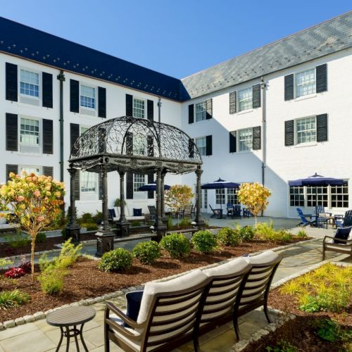 A bright hotel courtyard with white buildings, black shutters, a central gazebo, manicured gardens, and benches in a sunny, inviting setting.
