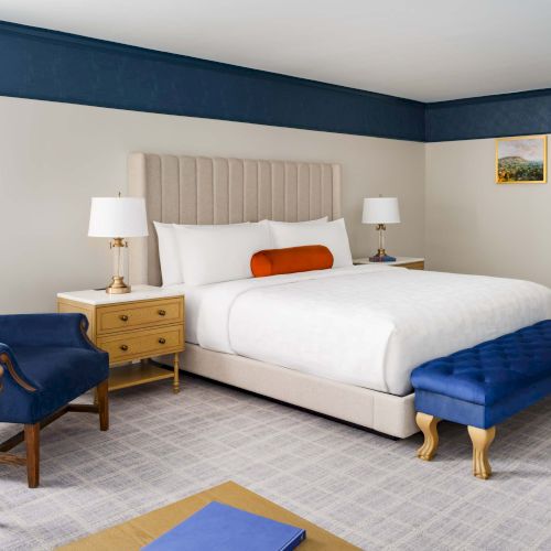 A bright hotel room with a large bed, blue accents, a bench at the foot, a pair of bedside tables, and a seating area with a blue chair and ottoman, ending with a cozy, tidy vibe.