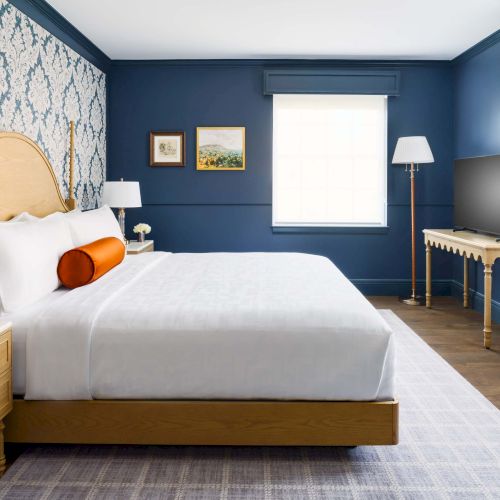 A stylish blue-toned hotel-style bedroom with a wooden bed, white linens, patterned accent wall, nightstands, lamps, a desk, and a flat-screen TV.