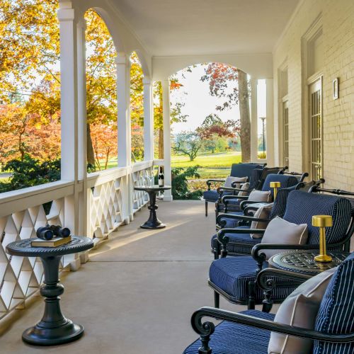 A covered porch with blue cushioned chairs, small tables, and autumn trees in the background, offering a cozy outdoor seating area.