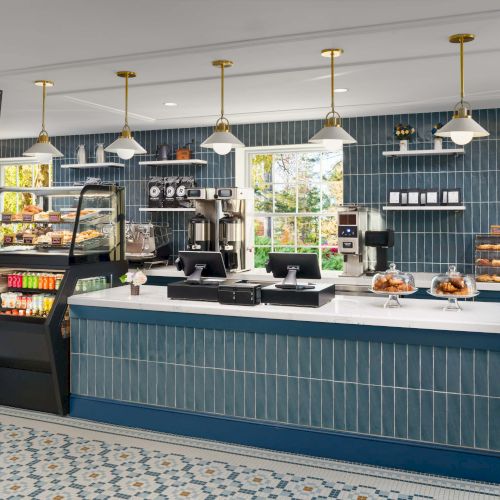 A modern cafe counter with blue tiled front, white countertop, pastry display, coffee machines, and a small menu board; warm pendant lights overhead.