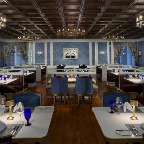 A stylish, upscale restaurant with blue tones, white tables, blue glassware, chandeliers, and cozy booths facing a framed wall art in a classic dining room.