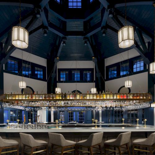 A stylish, dark-toned bar with a long counter, high chairs, hanging lanterns, and a vaulted ceiling; warm ambient lighting.