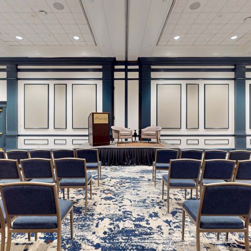 A conference room or small theater setup with rows of blue chairs facing a stage with a podium and a few items, in a carpeted blue-toned room.