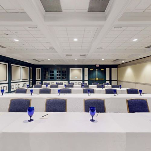 A bright conference room with long white tables, blue chairs, and blue-stemmed water glasses arranged in rows, ready for a meeting.