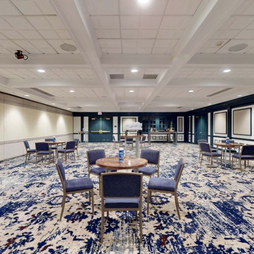 A spacious conference room with patterned blue carpet, round tables and chairs, wall panels, and a projector screen at the far end, blank and ready for an event.