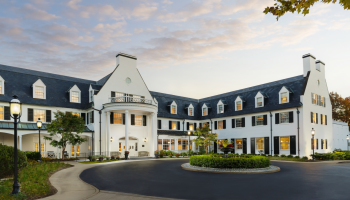 A white, colonial-style hotel with a U-shaped layout, neatly manicured entrance, roundabout, and warm lights at dusk.