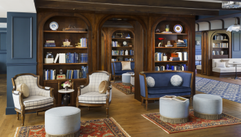 A luxurious, wood-paneled library lounge with blue seating, built-in bookshelves, cozy armchairs, ottomans, and oriental rugs in an elegant hotel-like setting.