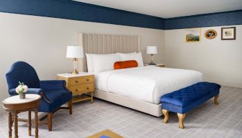 A bright hotel room with a large bed, blue accents, two nightstands, lamps, a blue armchair, a matching ottoman, and a small round table with flowers.