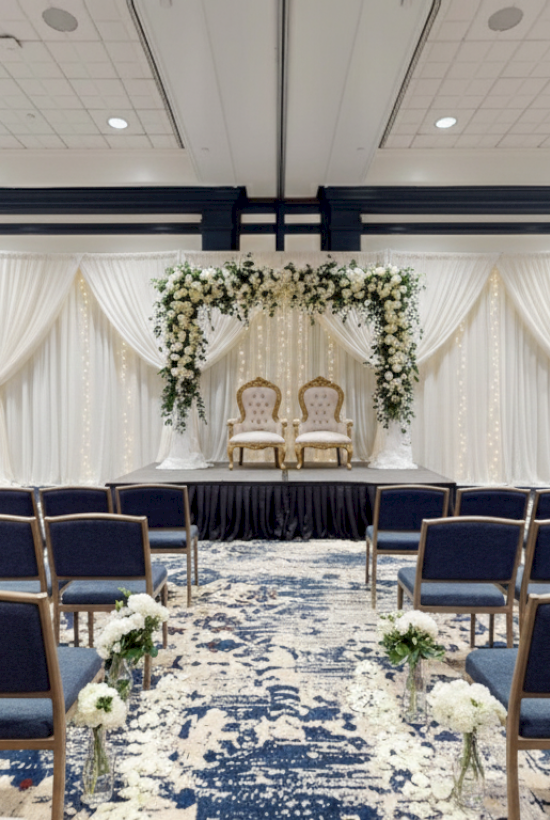 Wedding ceremony setup in a formal ballroom: a stage with white drapes, floral arch, two chairs, and rows of blue cushioned chairs facing it.