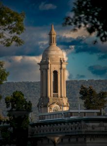 A tall, ornate stone monument or spire rises above a blue sky with clouds, framed by greenery and a decorative railing in the foreground.