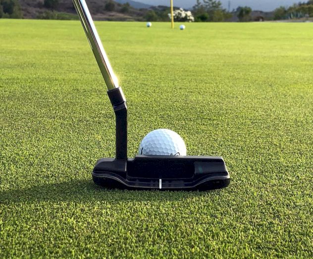 A golf ball rests on a black putter grip on a green putt surface, with a distant practice area in the background.