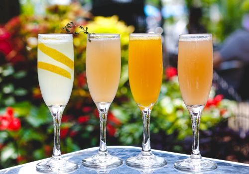Four elegant cocktails on a glass table with colorful flowers blurred in the background, each glass featuring a different fruity garnish.