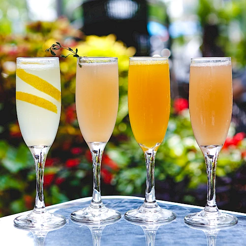 Four elegant cocktails on a glass table with colorful flowers blurred in the background, each glass featuring a different fruity garnish.