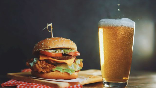 A juicy fried chicken burger with lettuce and tomato beside a frosty glass of beer, on a rustic wooden table.