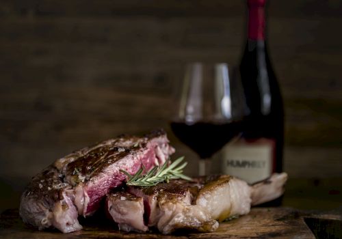 A plated grilled steak with rosemary on a wooden board, with a fork and knife blurred in the background.