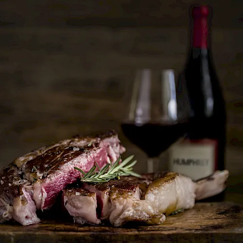 A plated grilled steak with rosemary on a wooden board, with a fork and knife blurred in the background.