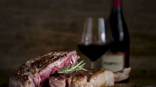 A plated grilled steak with rosemary on a wooden board, with a fork and knife blurred in the background.
