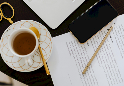 A coffee cup on a saucer with a wooden stirrer, beside a smartphone, scissors, laptop corner, and papers with a pencil on a dark desk.
