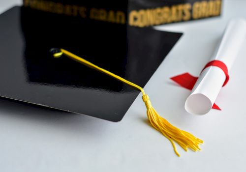 A graduation cap with a yellow tassel and a rolled diploma tied with a red ribbon.