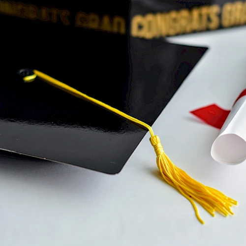 A graduation cap with a yellow tassel and a rolled diploma tied with a red ribbon.