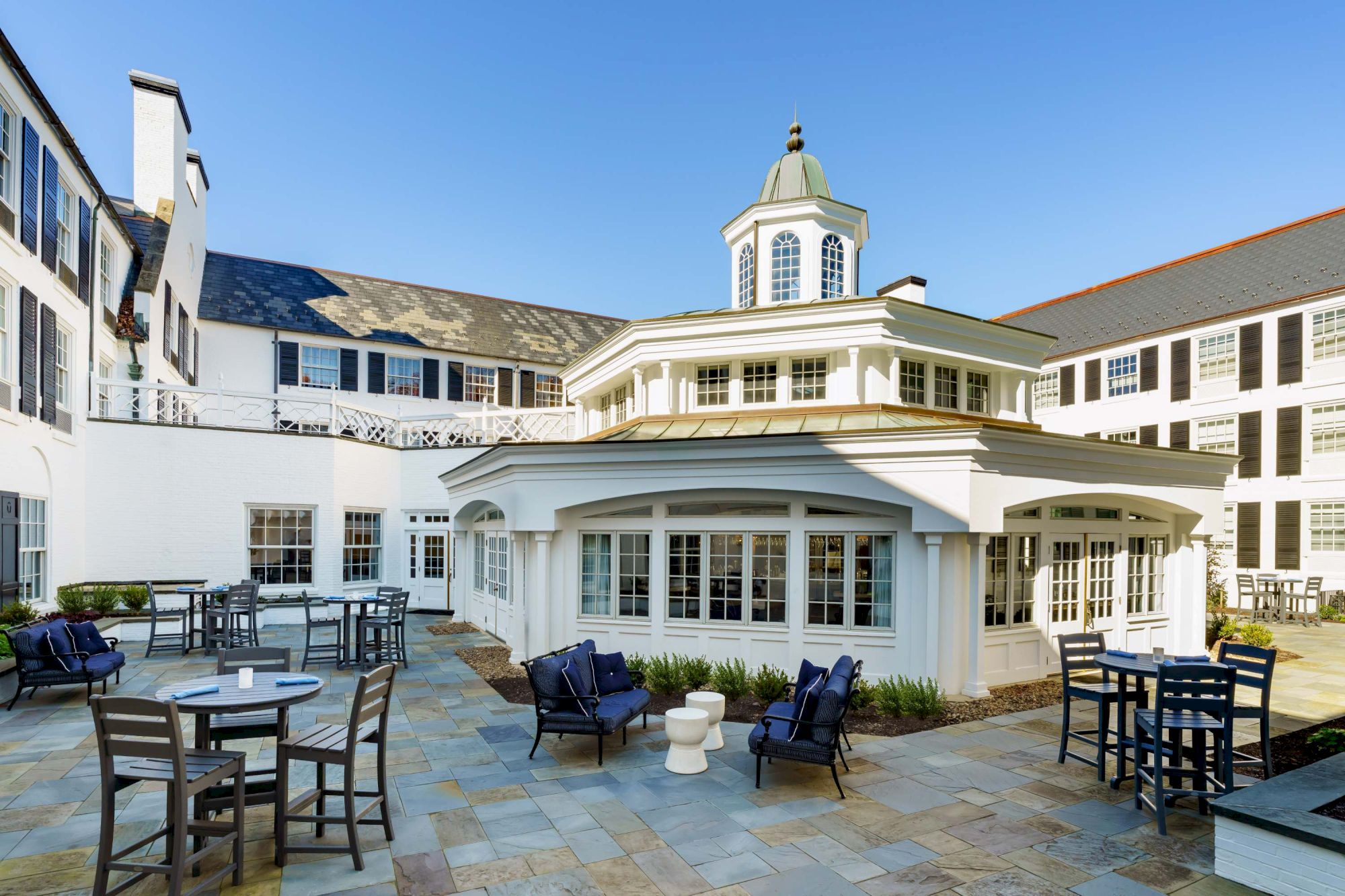 A bright hotel courtyard with a white, round, glass-fronted conservatory at its center, surrounded by outdoor tables and chairs under a clear blue sky.