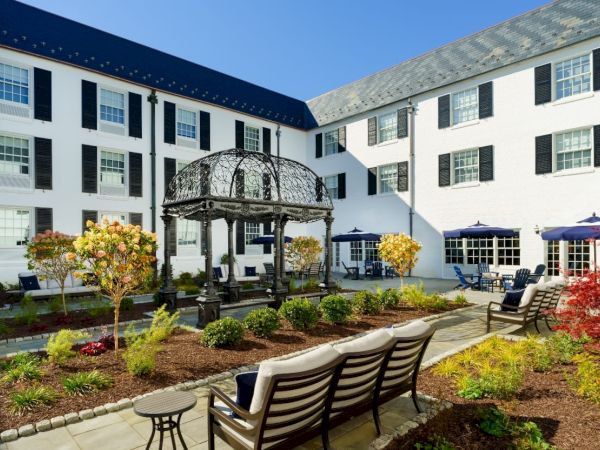 A hotel courtyard with white-faced building, black shutters, garden beds, benches, and a decorative gazebo in the center, sunny day.