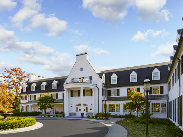A large white-and-gray hotel with a curved driveway, manicured landscaping, and a bright sky; classic colonial-style building with dormer windows.