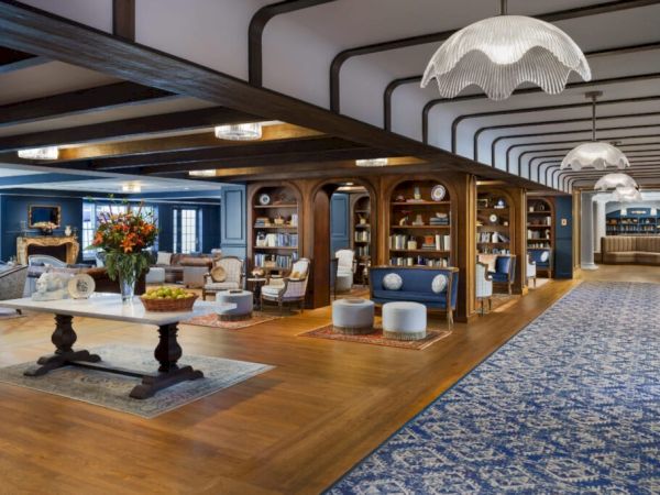 A spacious, elegant lounge with wooden beams, comfortable seating, bookshelves, tables, and large blue carpeted areas, plus decorative lantern lights.