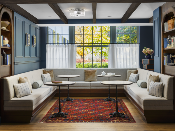 A cozy corner cafe nook with blue wall panels, built-in white sofas, small round tables, a red patterned rug, and large windows with sheer curtains.