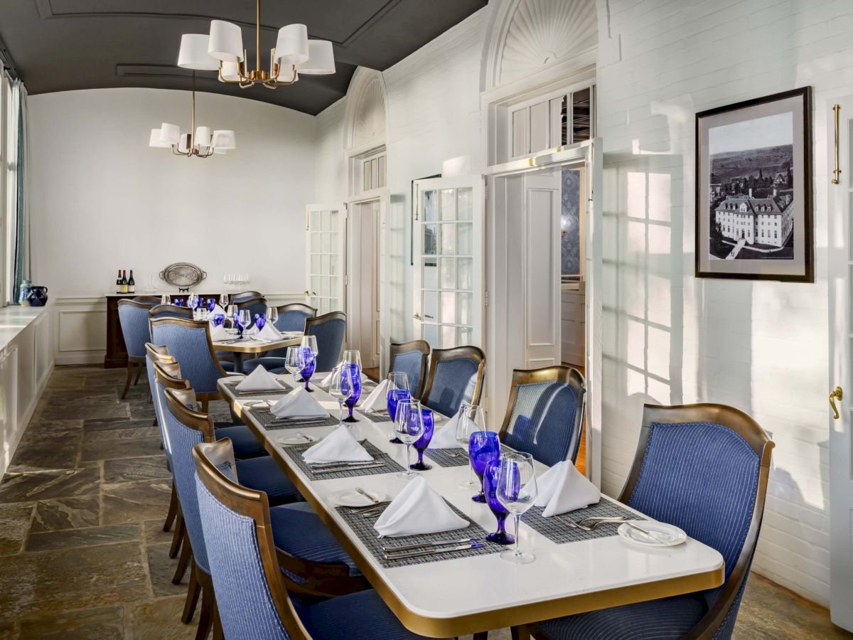 A long dining table set for a formal meal in a bright, elegant room with blue upholstered chairs, white napkins, blue glasses, and framed art on the wall. The table is neatly arranged with plates and cutlery, ready for guests, and soft natural light fills the space.