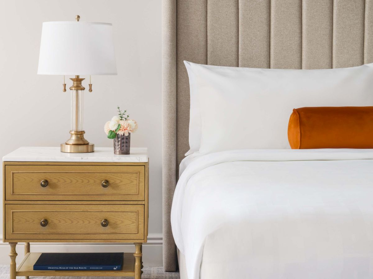 A cozy bedroom scene with a nightstand, brass lamp, small vase of flowers, and a neatly made bed with a white duvet and an orange bolster.