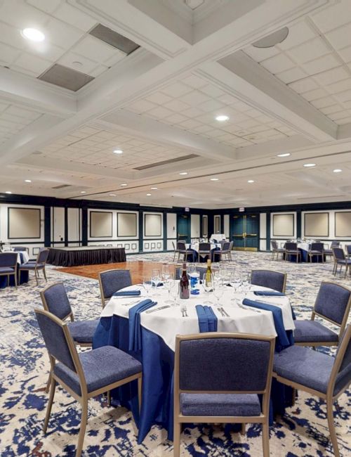 A large banquet hall with round tables draped in blue and white linens, blue chairs, and a patterned carpet, set for an event.