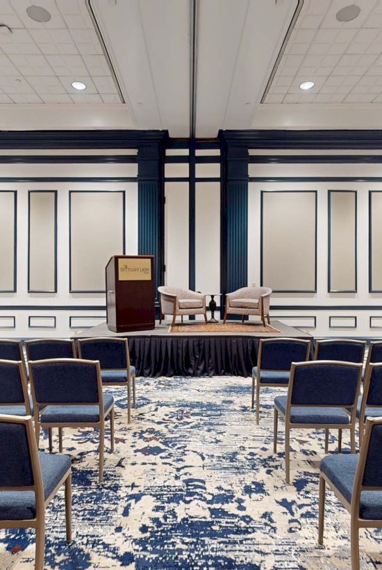 A conference room set for a panel or TED-style talk: podium, two chairs, backdrop panels, and rows of navy chairs facing the front.