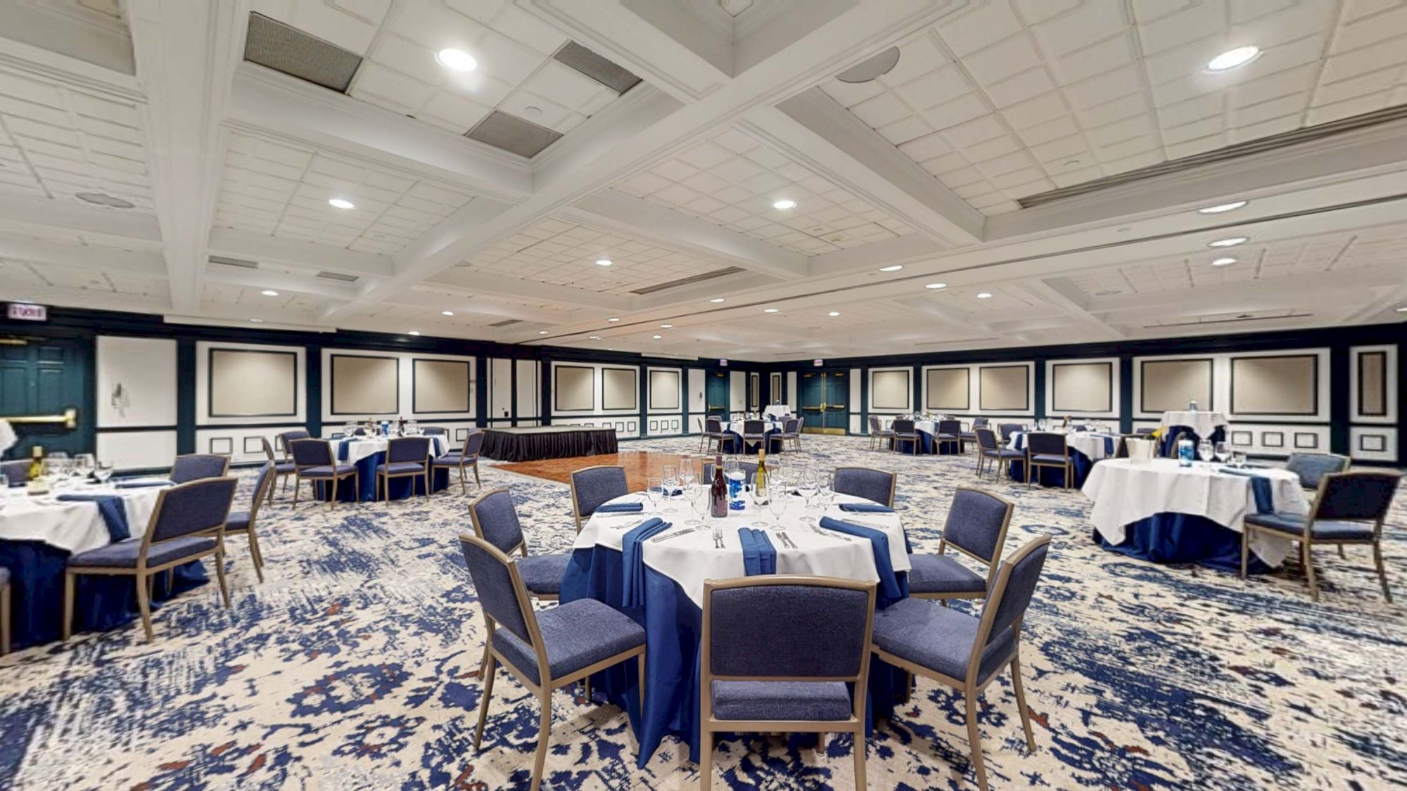 A large banquet hall set for a formal event, round tables with blue tablecloths and chairs, chandeliers, and decorative wall panels.