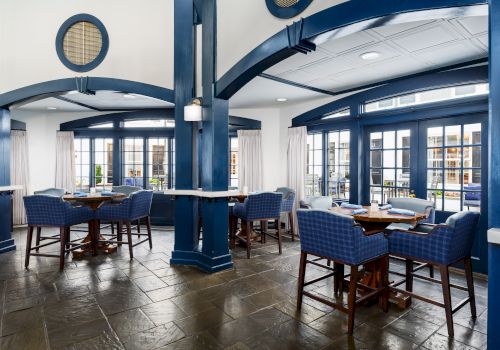 Bright modern dining lounge with blue columns, wood tables, upholstered chairs, white curtains, and large glass doors and windows.