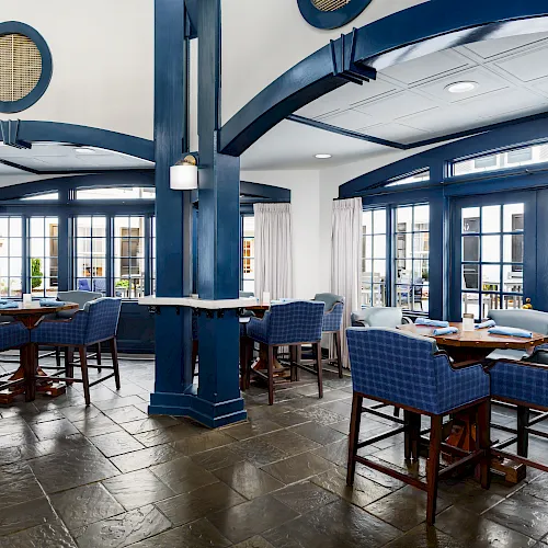 Bright modern dining lounge with blue columns, wood tables, upholstered chairs, white curtains, and large glass doors and windows.