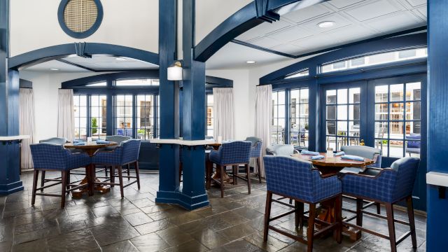 Bright modern dining lounge with blue columns, wood tables, upholstered chairs, white curtains, and large glass doors and windows.