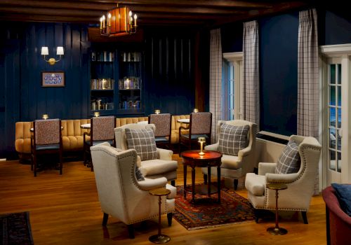 An elegant lounge with upholstered chairs, a glowing table lamp, tall windows with curtains, and a bookshelf in a cozy blue room.
