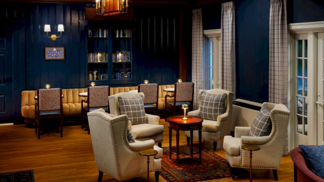 An elegant lounge with upholstered chairs, a glowing table lamp, tall windows with curtains, and a bookshelf in a cozy blue room.