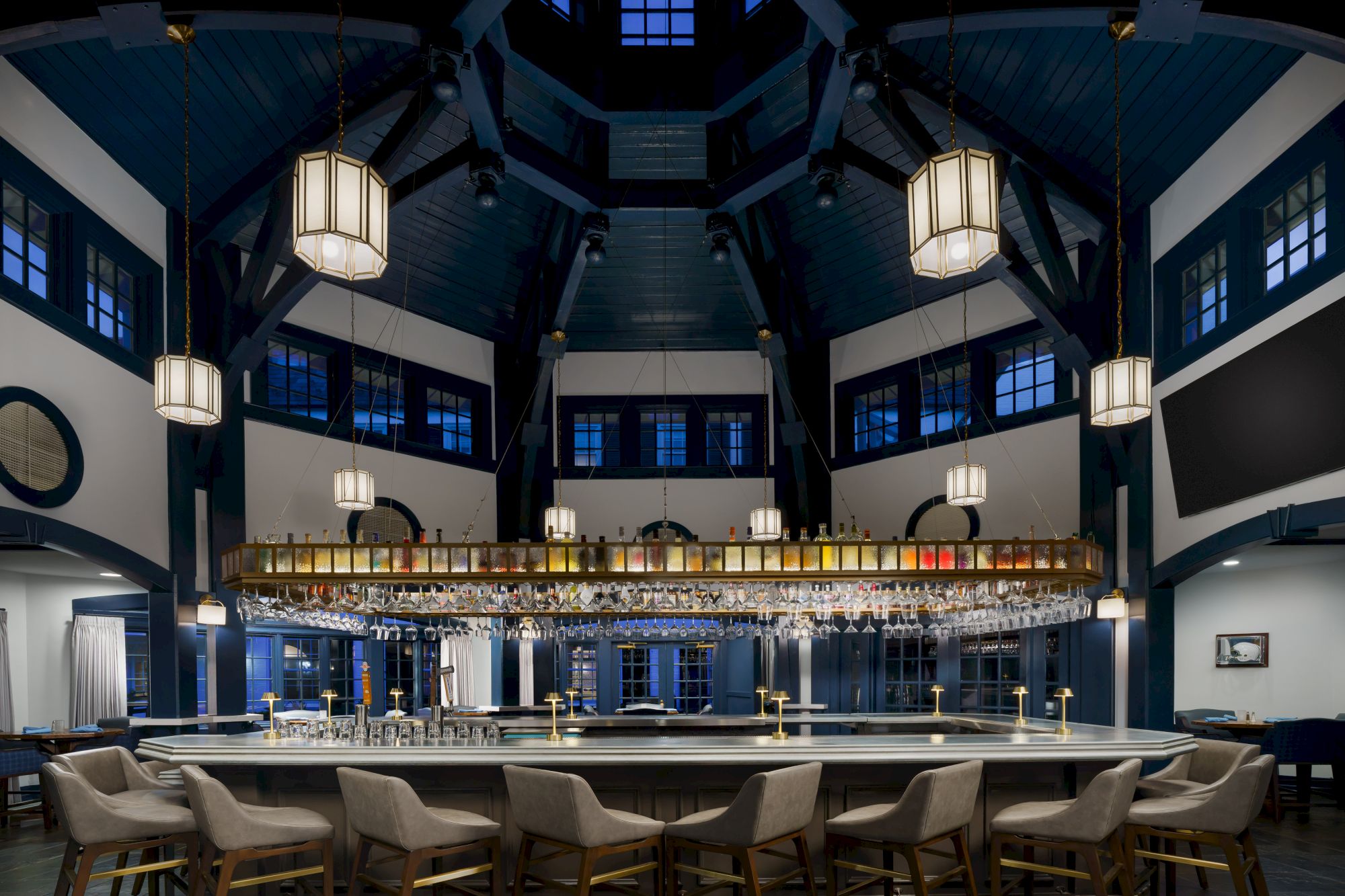 A chic bar interior with hanging lantern lights, stocked bottles and glassware, and rows of stools under a high vaulted ceiling.
