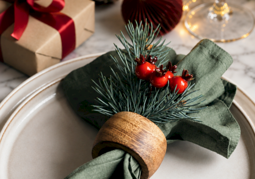 A holiday table setting with a green napkin, pine branches, red baubles, a pinecone, wrapped gifts, and warm string lights.