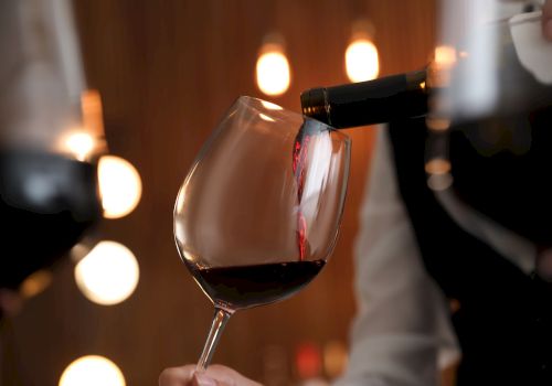 A bottle pours red wine into a stemmed glass, with warm bokeh lights glowing in the background.